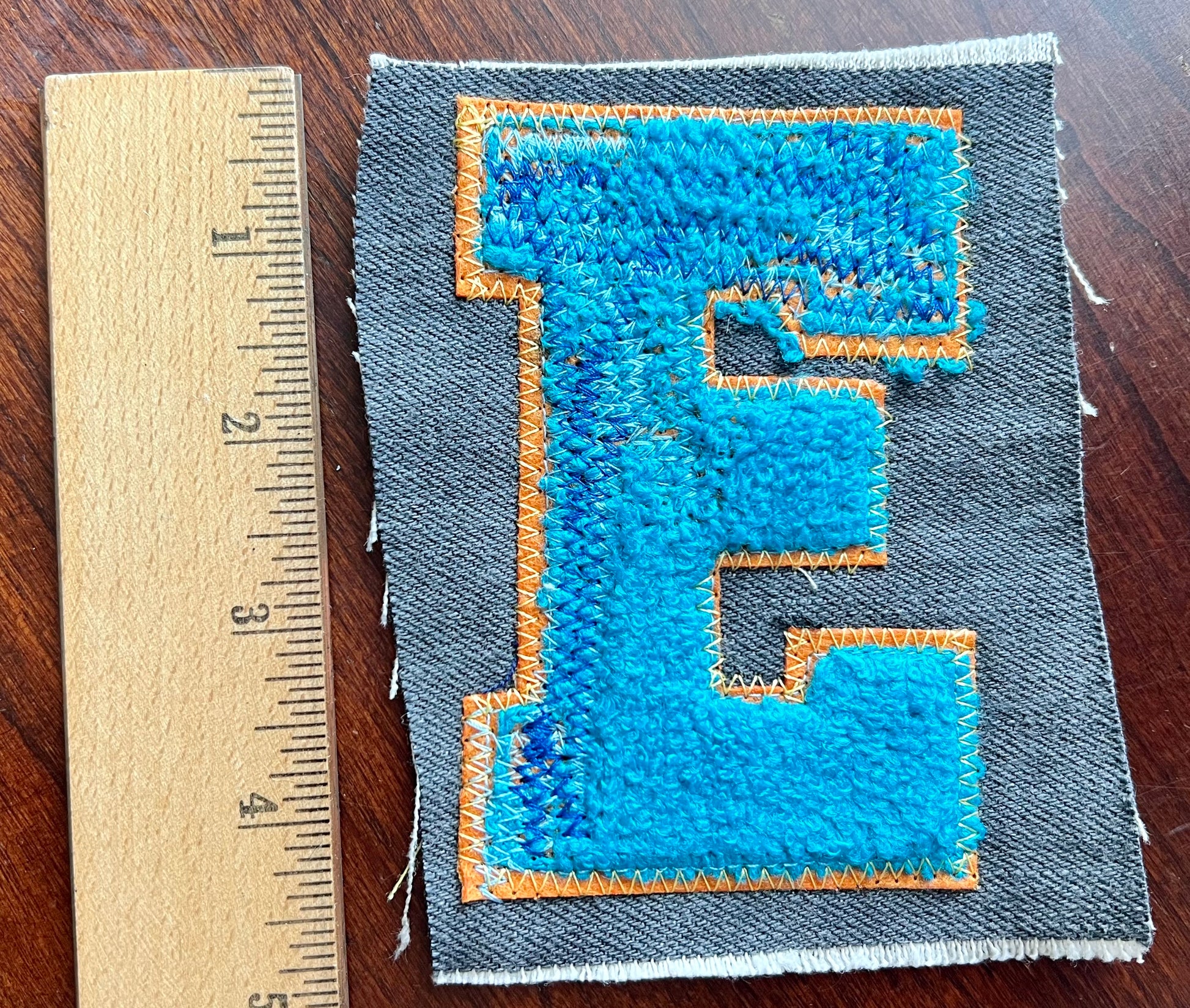 Fabric patch with blue letter 'E' on gray background, next to a ruler for scale.