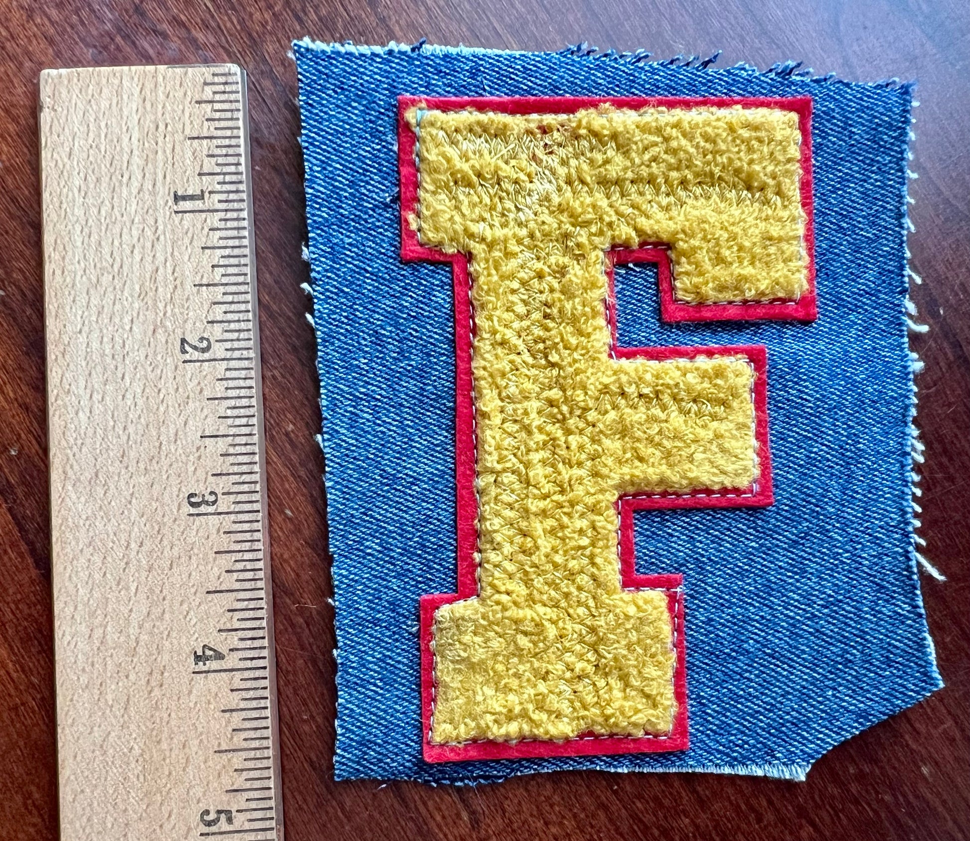 Letter 'F' patch on denim fabric with a ruler for scale on a wooden surface
