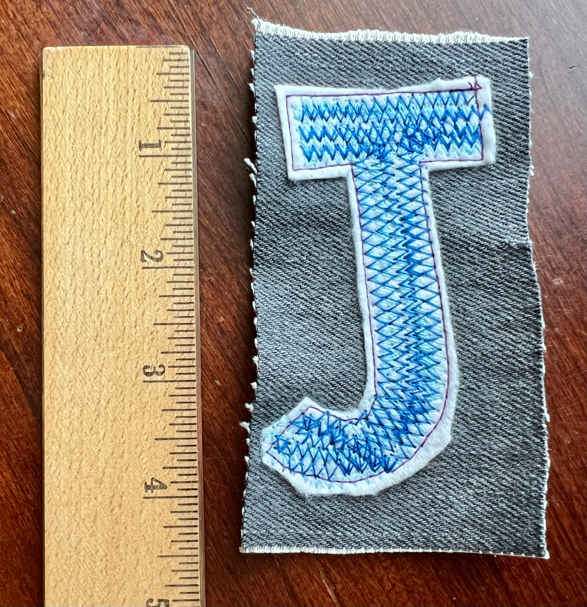 Denim patch with blue embroidered letter 'J' next to a wooden ruler on a wooden surface