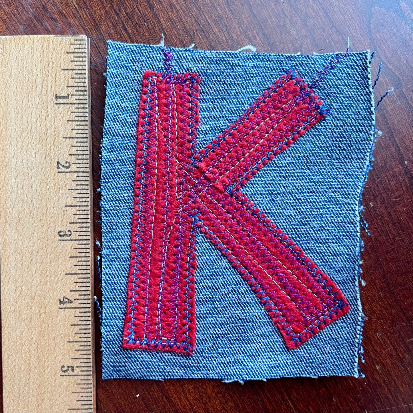 Fabric patch with red letter 'K' on a wooden surface with a ruler for scale.