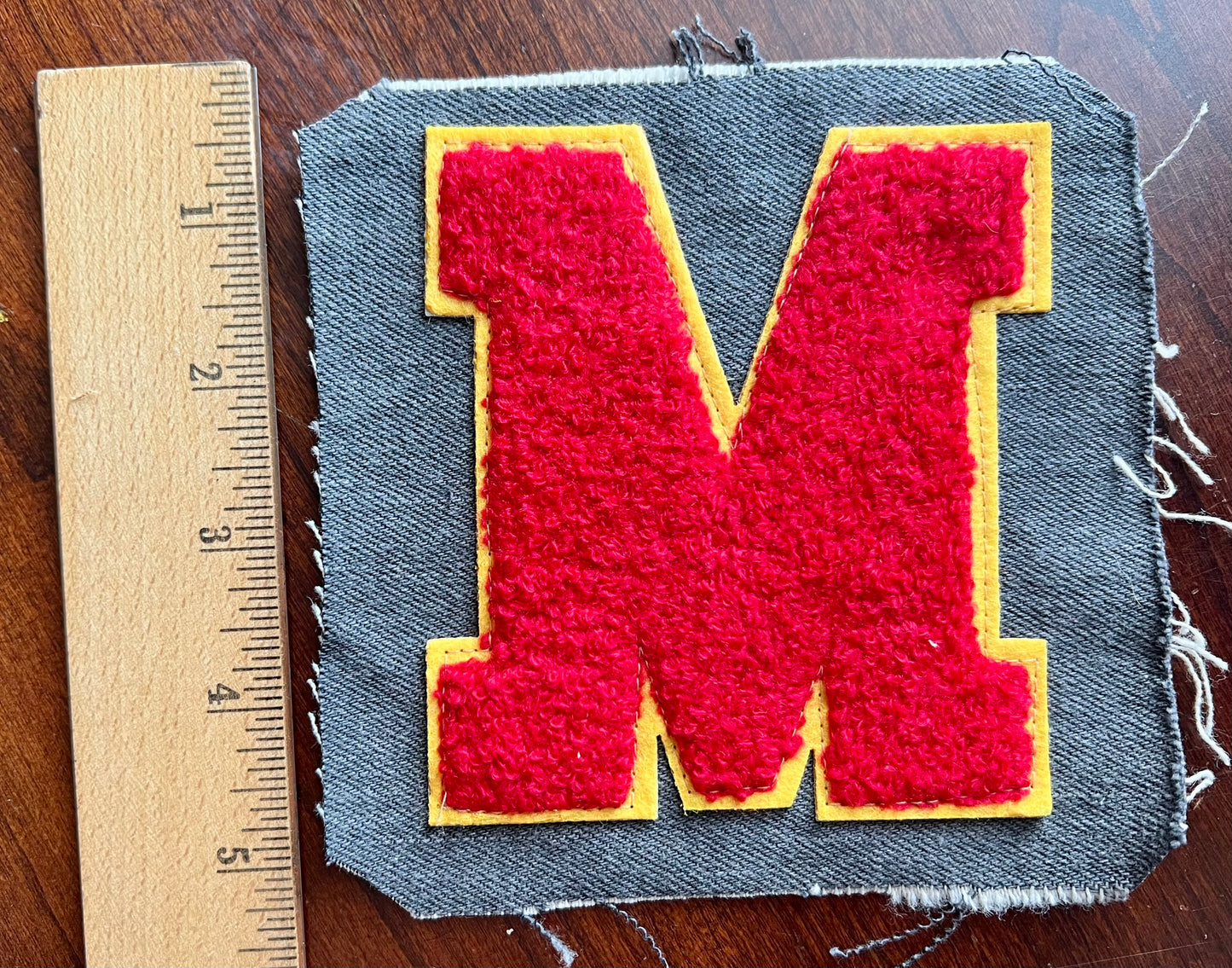 Red letter 'M' patch on gray fabric with yellow border, next to a ruler for scale.