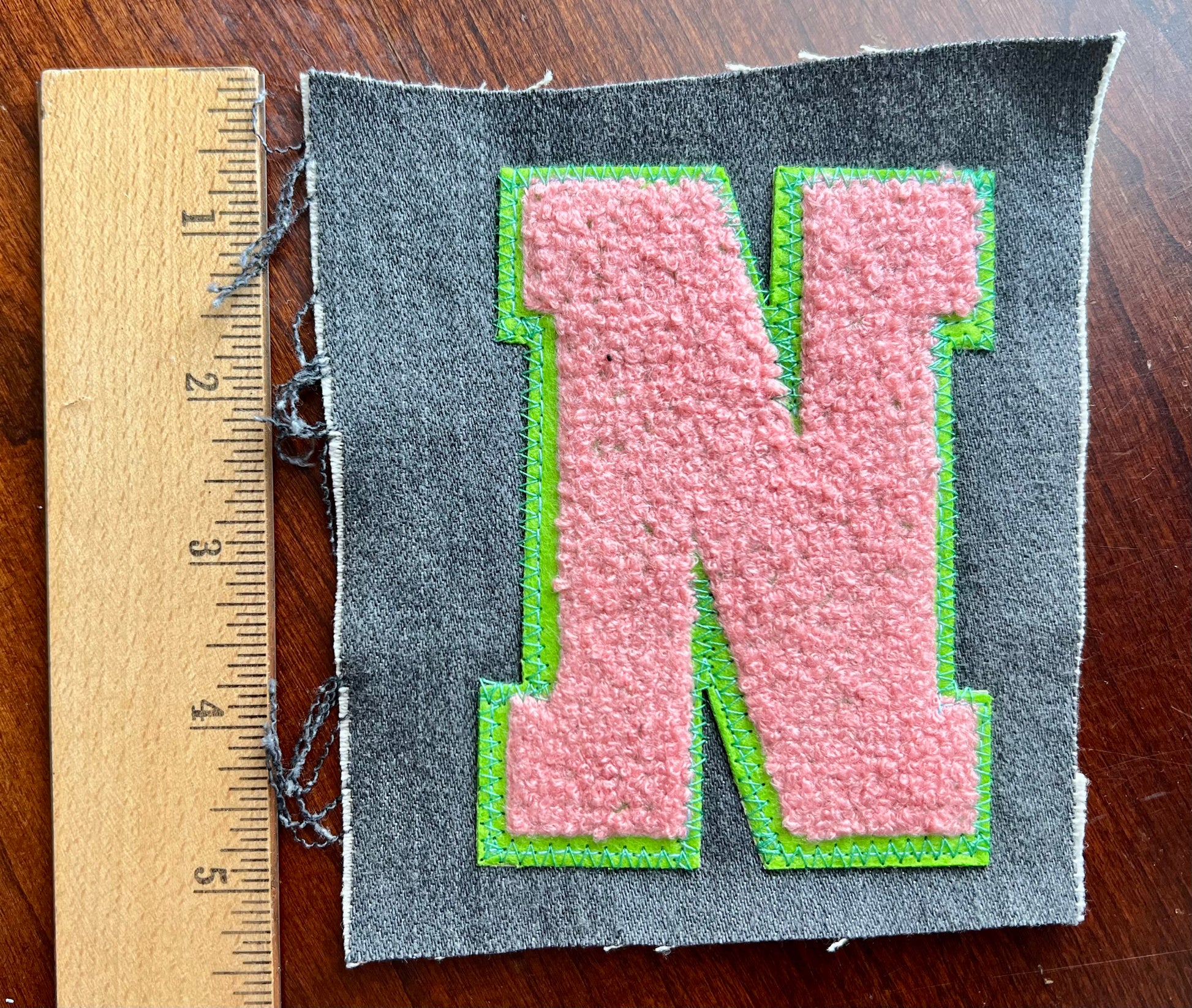 Fabric patch with a pink letter 'N' on gray fabric, next to a wooden ruler for scale.