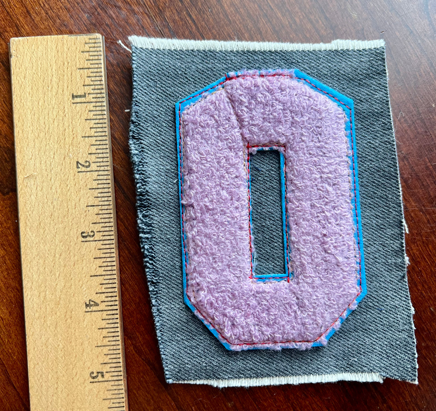 Fabric patch with a purple emblem on gray fabric, next to a ruler for scale, on a wooden surface.