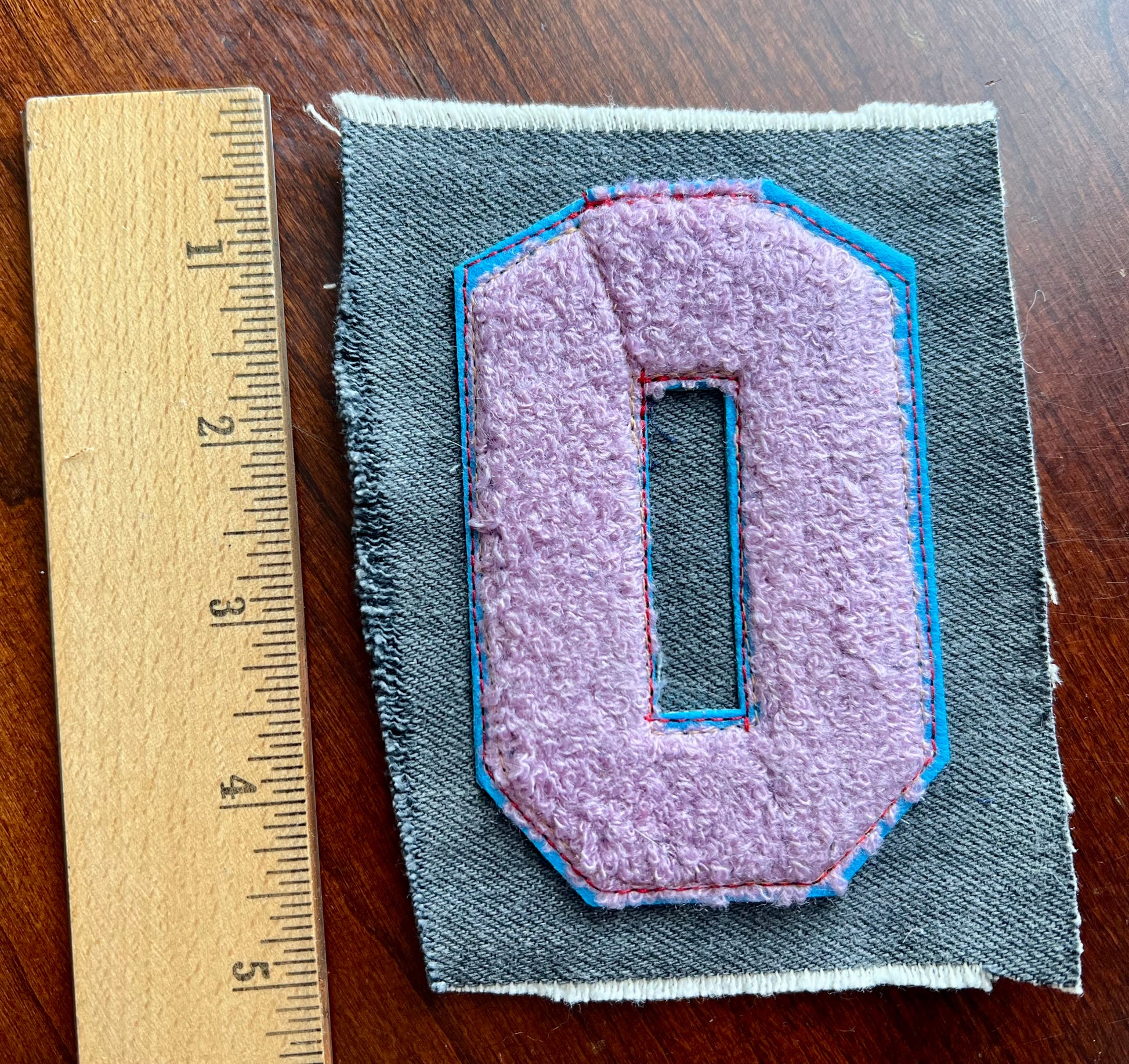 Fabric patch with a purple emblem on gray fabric, next to a ruler for scale, on a wooden surface.