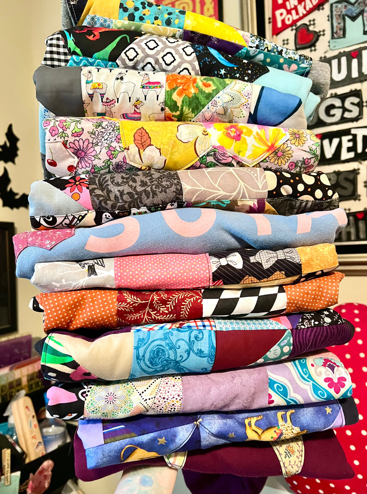 A tall stack of folded quilted sweatshirts with a variety of colorful floral, geometric, and patterned fabrics, photographed in a home setting with framed wall art in the background.