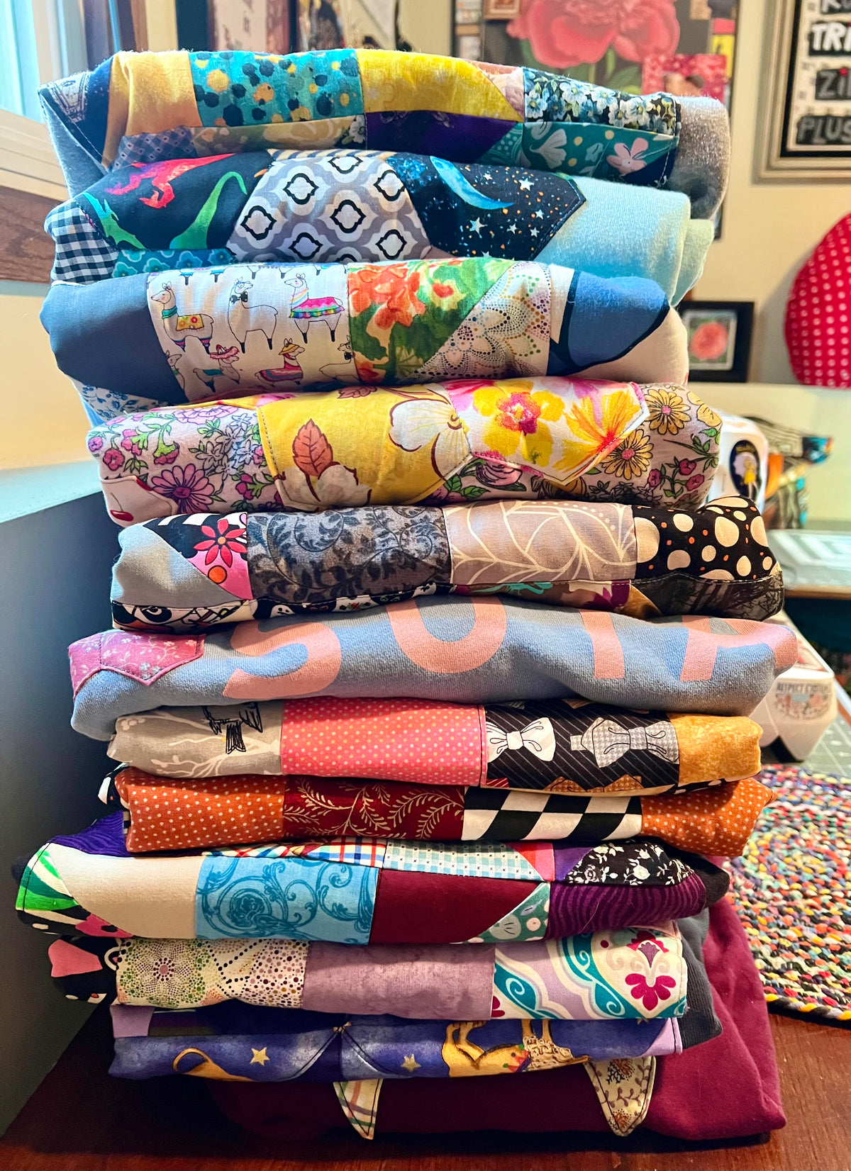 A tall stack of folded quilted sweatshirts with various colorful patchwork patterns, including florals, polka dots, stripes, and geometric designs, photographed in a home setting.