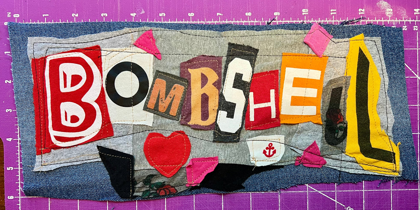 Textile art piece with colorful letters spelling 'BOMBSHELL' on a purple cutting mat.