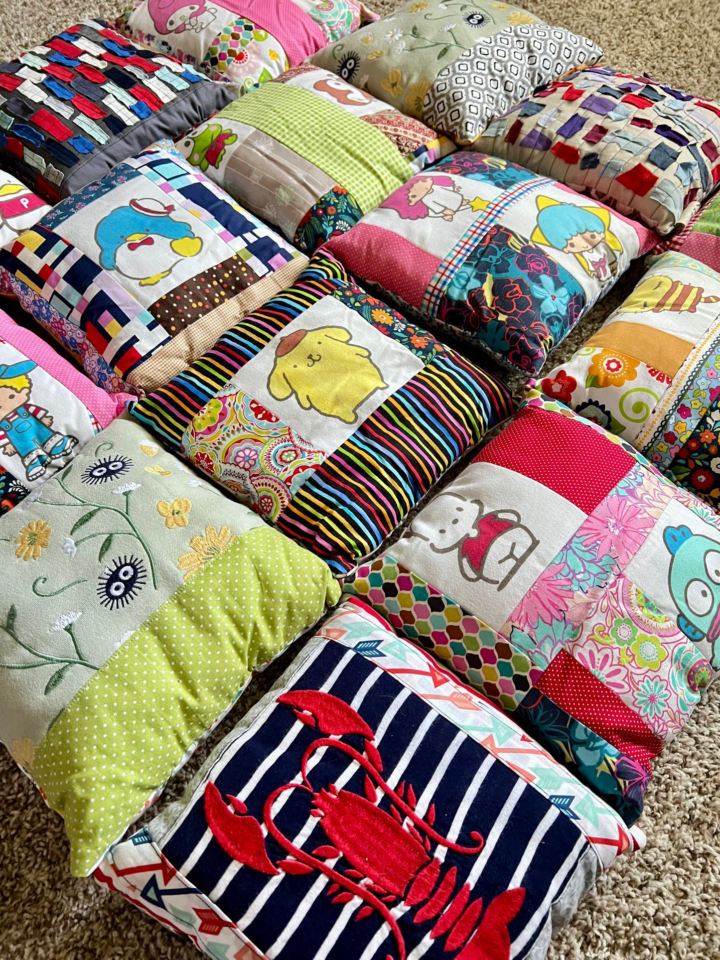 A collection of colorful handmade patchwork pillows arranged on a beige carpet. The pillows feature various patterns including stripes, florals, cartoon characters, and a red lobster design.