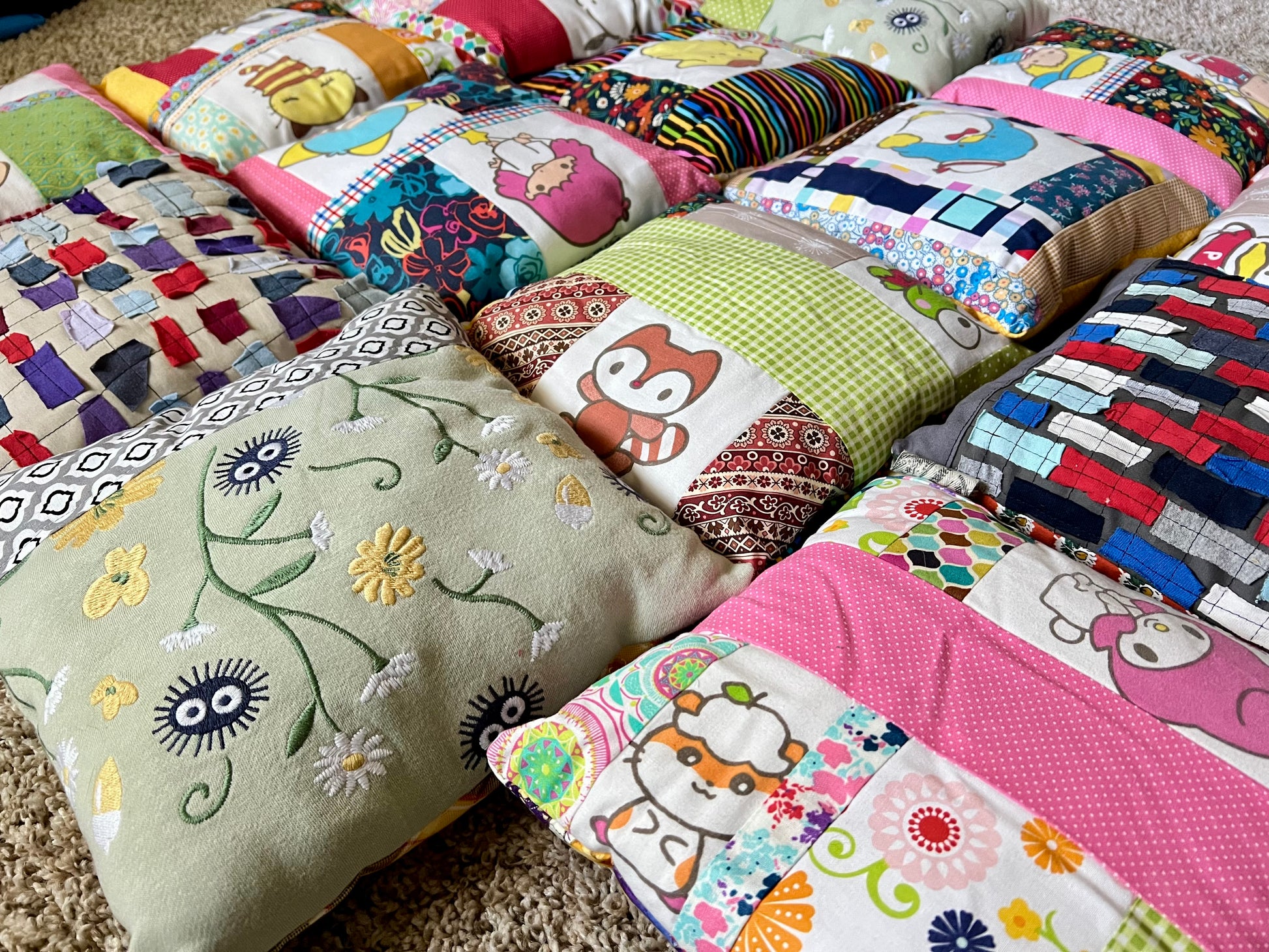 A collection of handmade, colorful patchwork pillows arranged on a carpeted floor. The quilts feature various patterns including floral embroidery, cartoon characters, stripes, and geometric designs in pink, green, blue, yellow, and red.