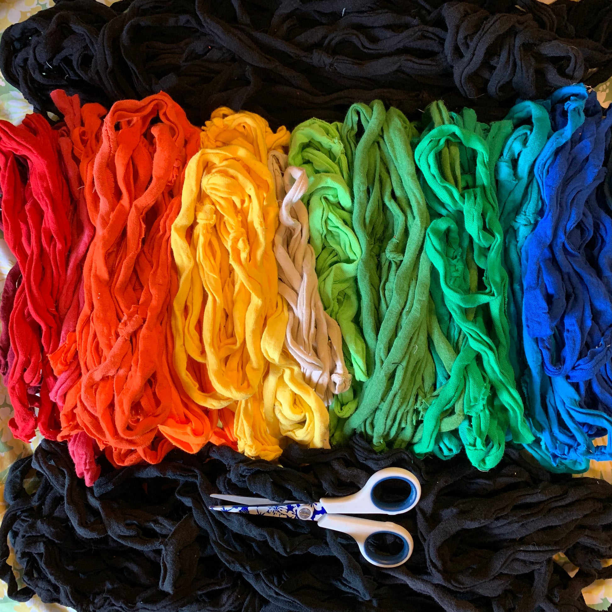 A flat-lay arrangement of colorful t-shirt scraps/ strips in a rainbow sequence, including red, orange, yellow, off-white, light green, medium green, and blue, with black scraps piled around and a pair of scissors at the bottom.