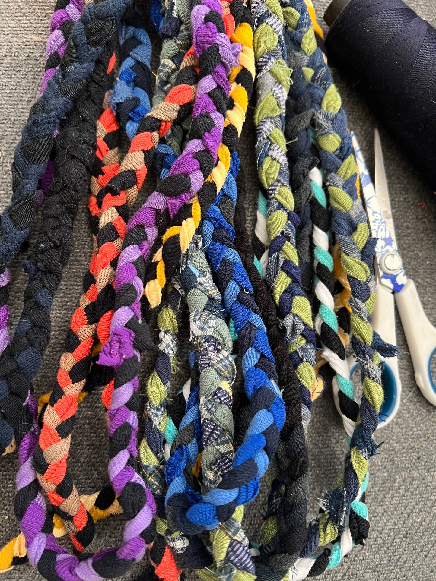 A close-up of several braided ropes made from mixed colorful fabric scraps, arranged on a gray textured surface with scissors and a spool of black rope partially visible.