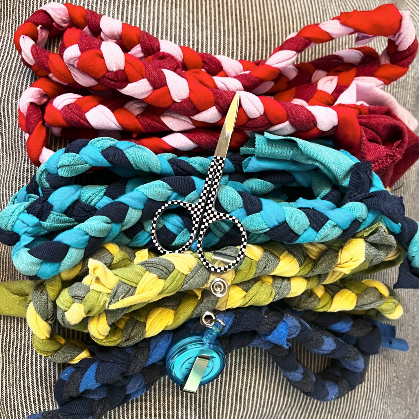 A stack of colorful braided fabric ropes in red, pink, turquoise, navy, yellow, and olive tones, with a pair of scissors and a blue hanging charm placed on top, set against a striped fabric background.
