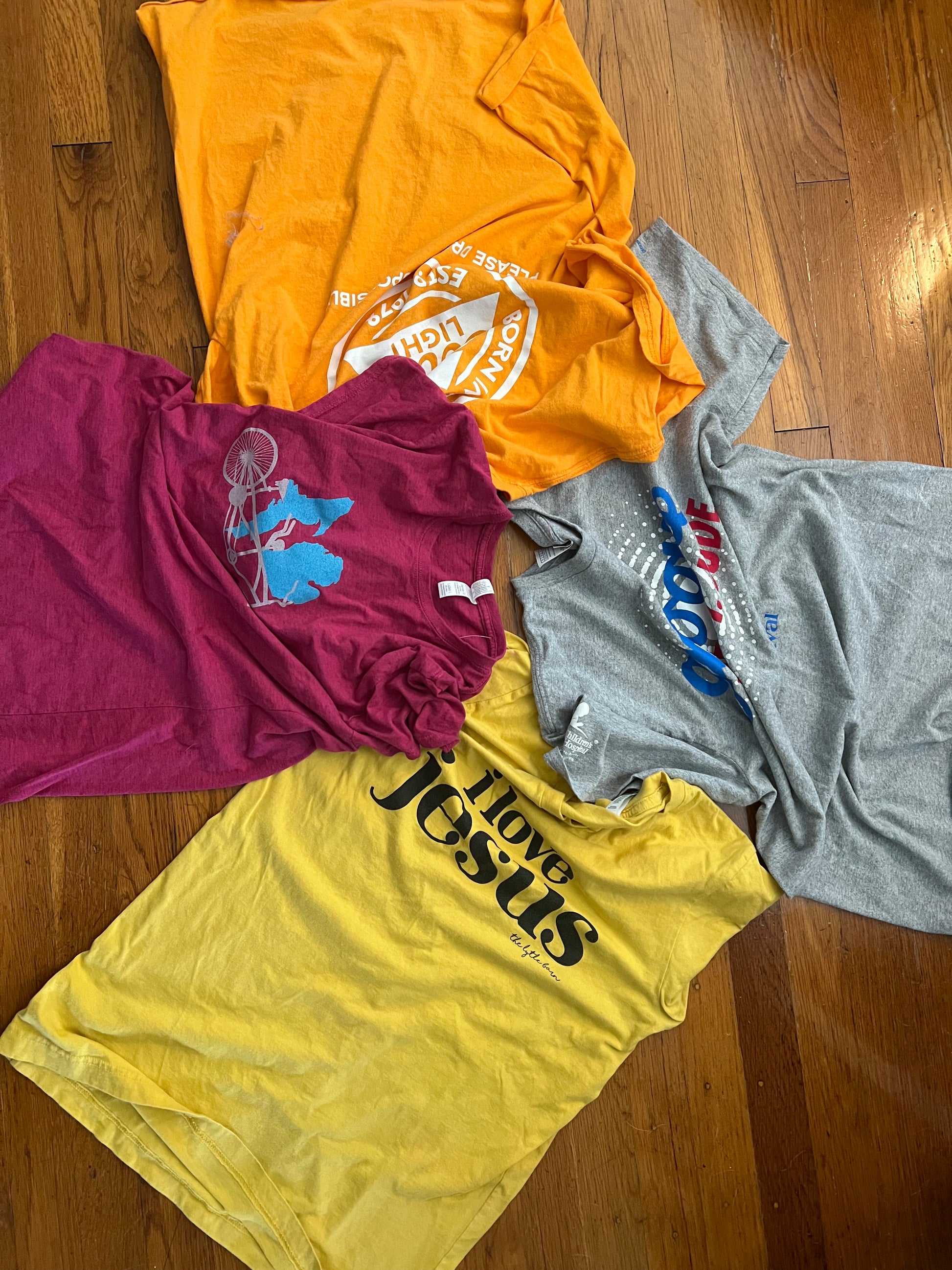 Four vintage graphic t-shirts in yellow, orange, magenta, gray, and light blue, arranged in a circle on a wooden floor. The shirts feature various words and images.