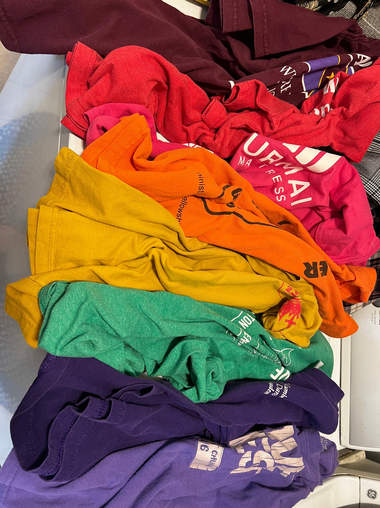 A pile of folded colorful t-shirts in various shades including purple, green, yellow, orange, red, and maroon, stacked on a light-colored surface.