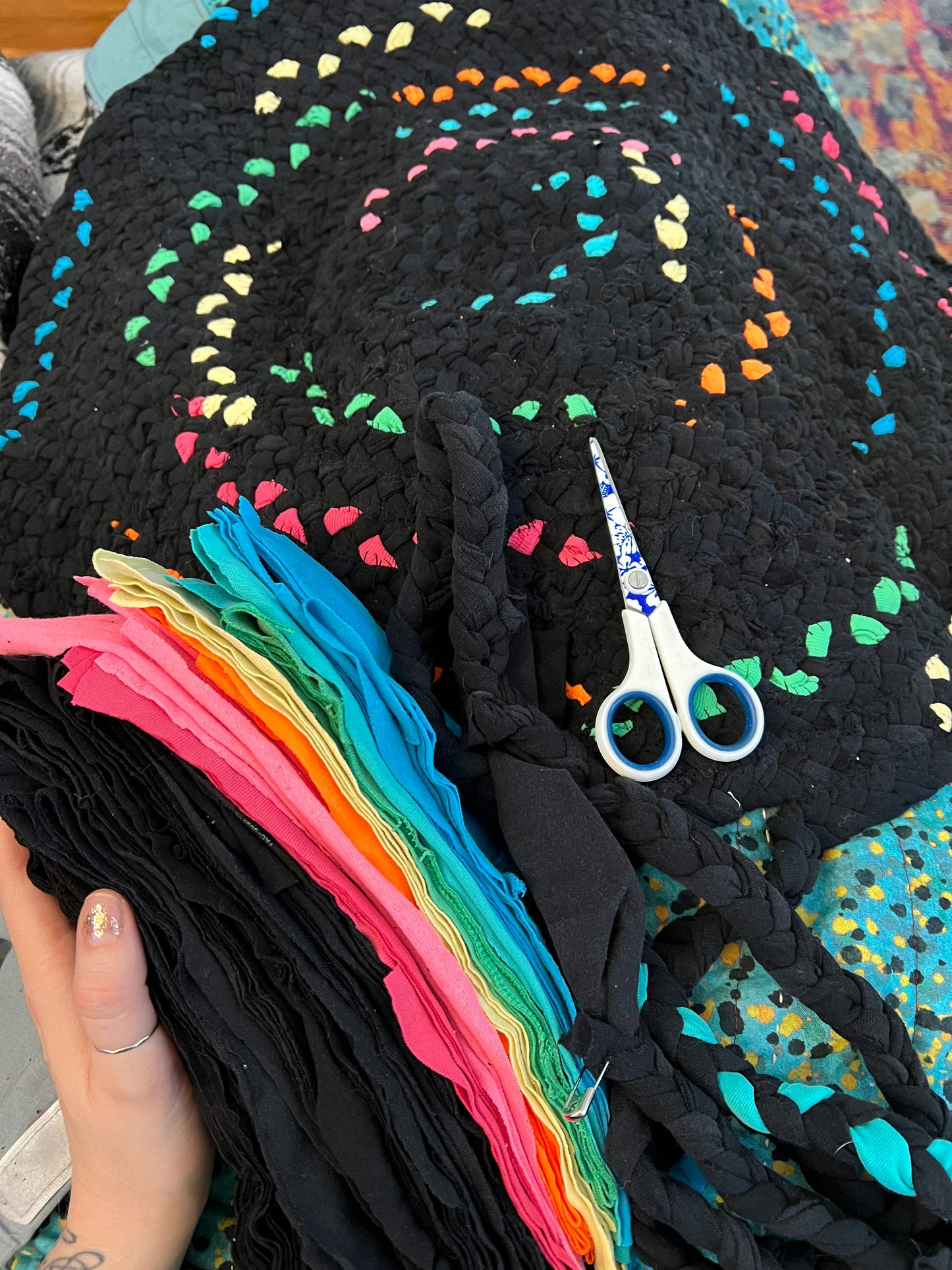 A close-up of a handmade black braided rug. A hand holds a stack of black fabric strips on the left, and a pair of blue-handled scissors rests on the braid.