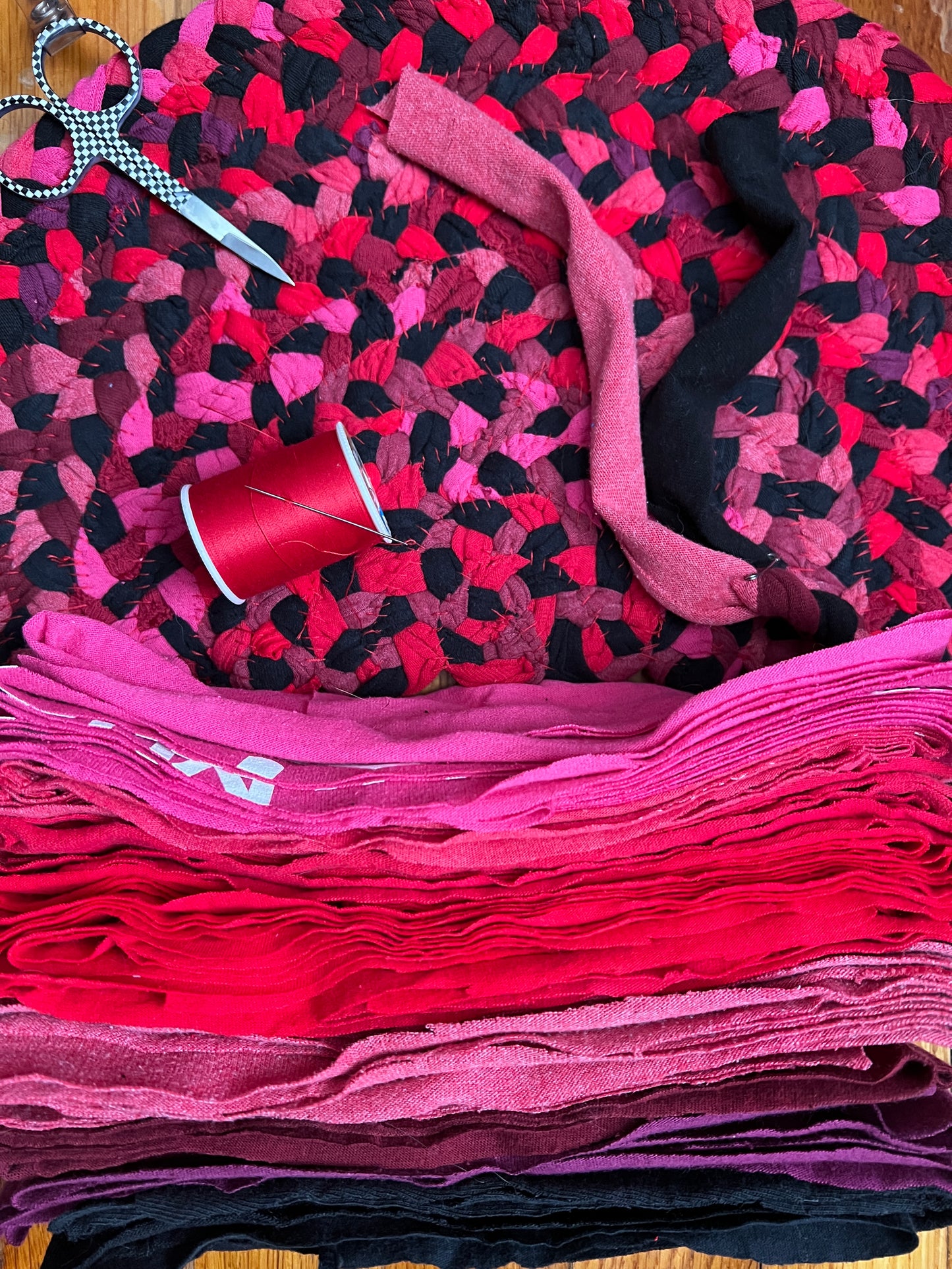A stack of layered fabric strips in bright pink, red, magenta, and black, arranged on a matching patterned fabric surface with a spool of red thread and scissors.