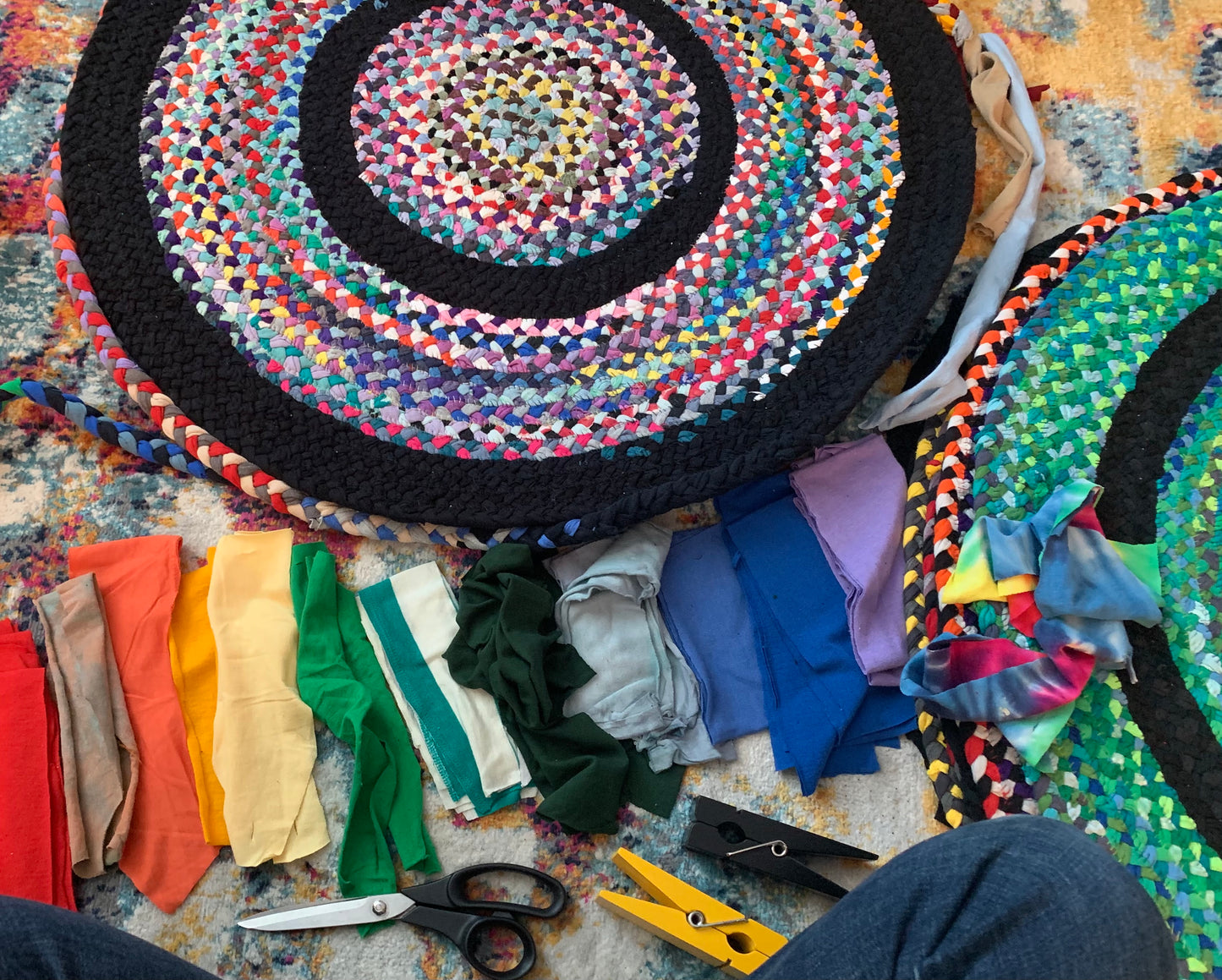 A top-down view of handmade braided rug pieces in progress, featuring a large circular rug with concentric rings of black and multicolor braided yarn. Scissors, a yellow utility tool, and folded strips of colorful fabric are arranged on a patterned rug, with a person's legs visible in the bottom corner.