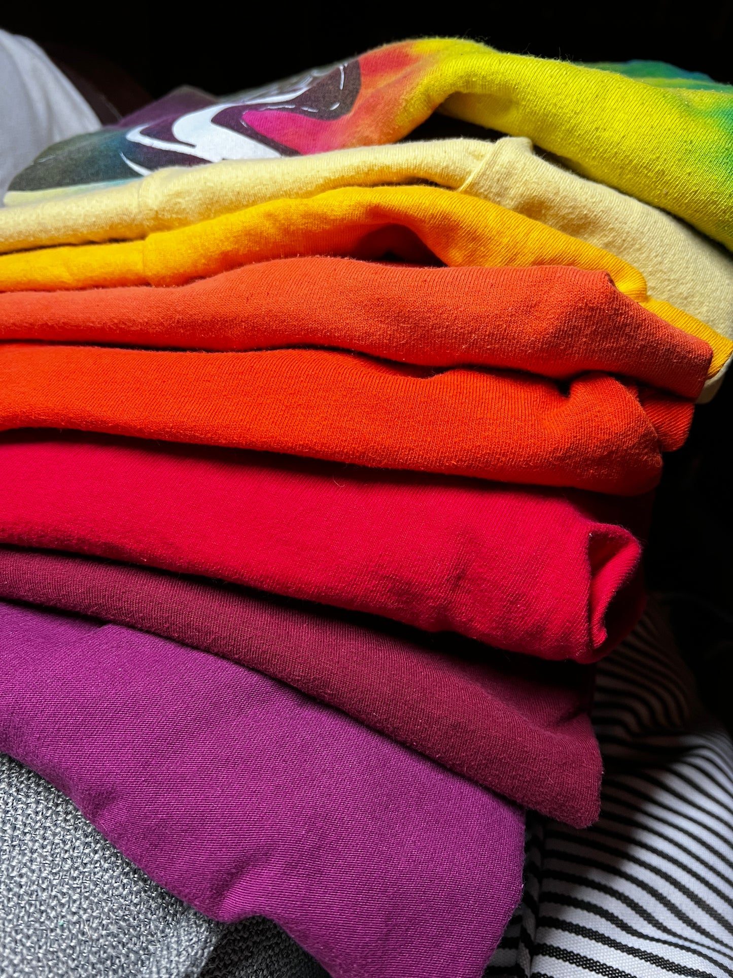 A close-up stack of folded colorful t-shirts in a rainbow gradient, showing shades of yellow, orange, red, pink, and purple.