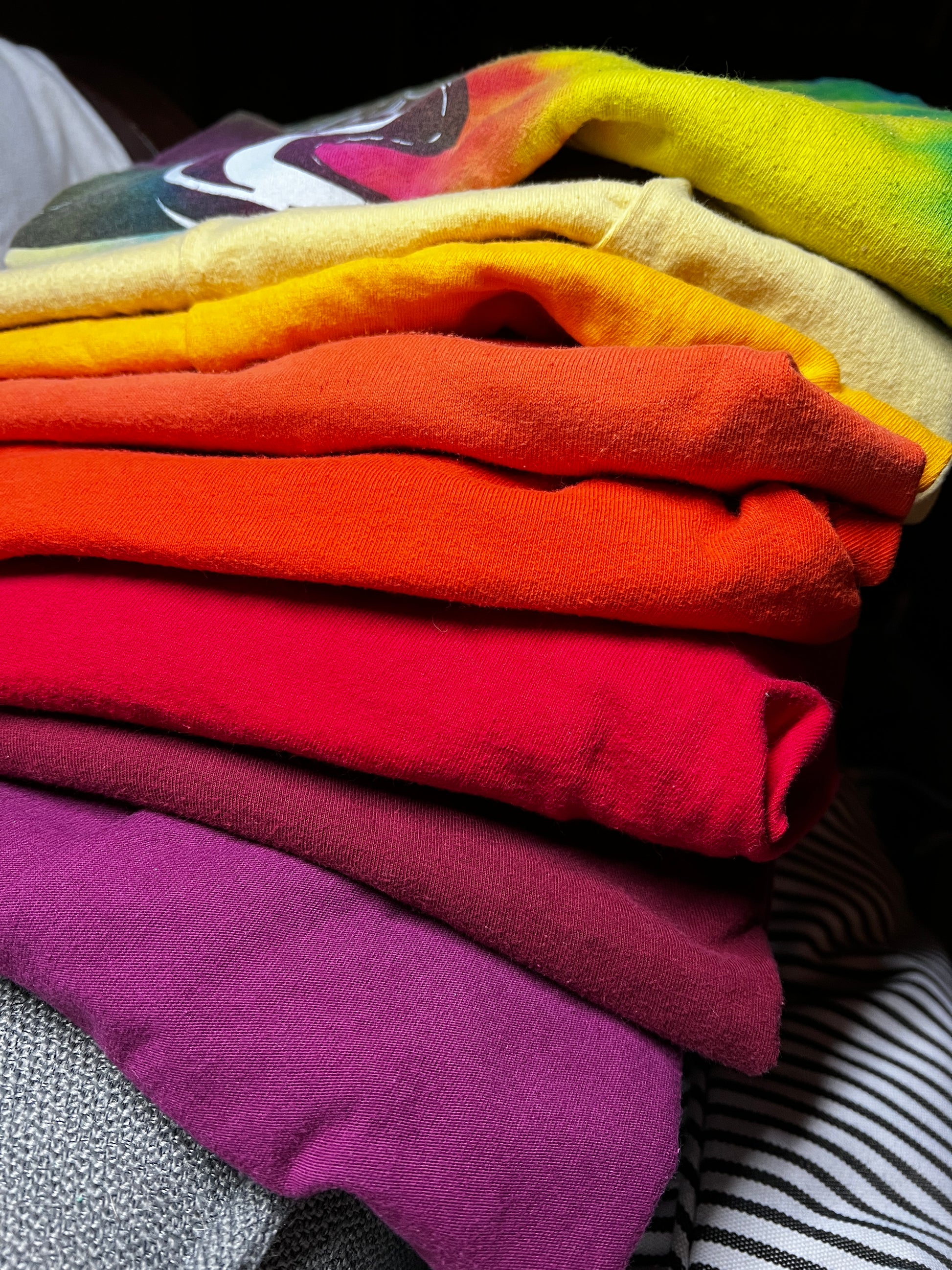 A close-up stack of folded colorful t-shirts in a rainbow gradient, showing shades of yellow, orange, red, pink, and purple.