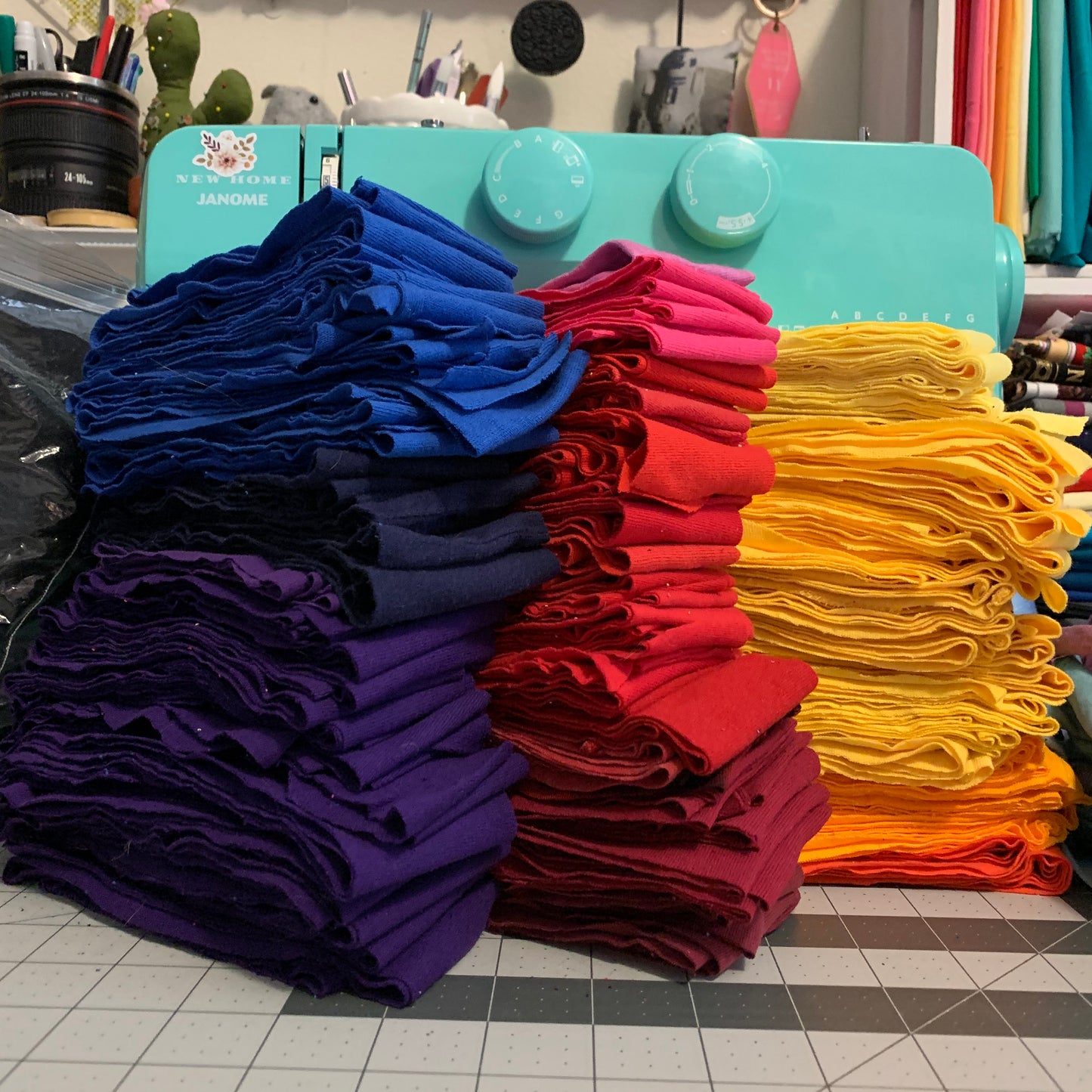 Three large stacks of folded fabric tshirt strips in blue, red, and yellow are arranged in front of a turquoise sewing machine on a tiled craft table.