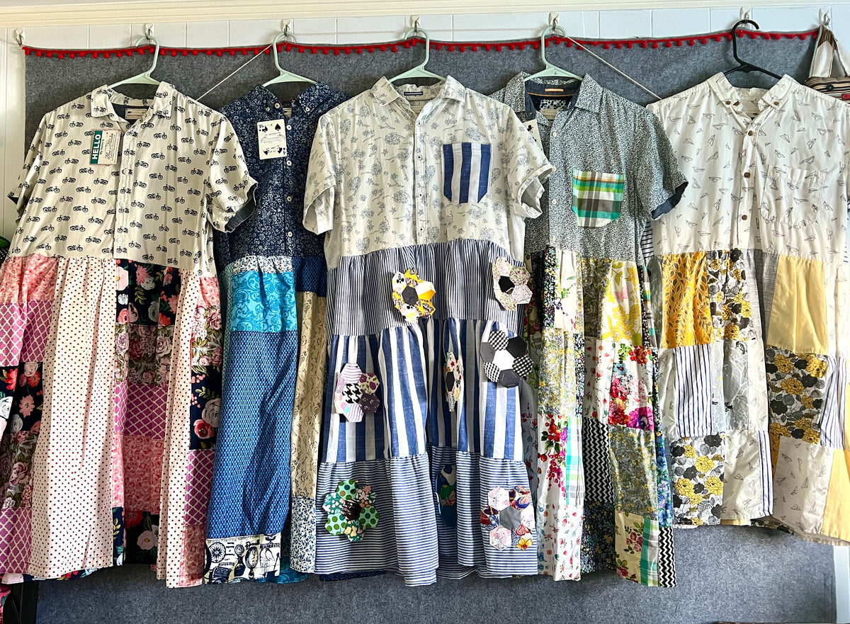 A collection of dresses made from patchwork fabrics and reclaimed button-up shirts, featuring various patterns and colors, displayed on hangers.
