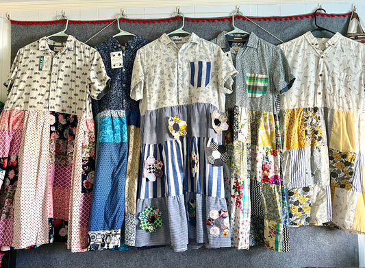 A collection of dresses made from patchwork fabrics and reclaimed button-up shirts, featuring various patterns and colors, displayed on hangers.