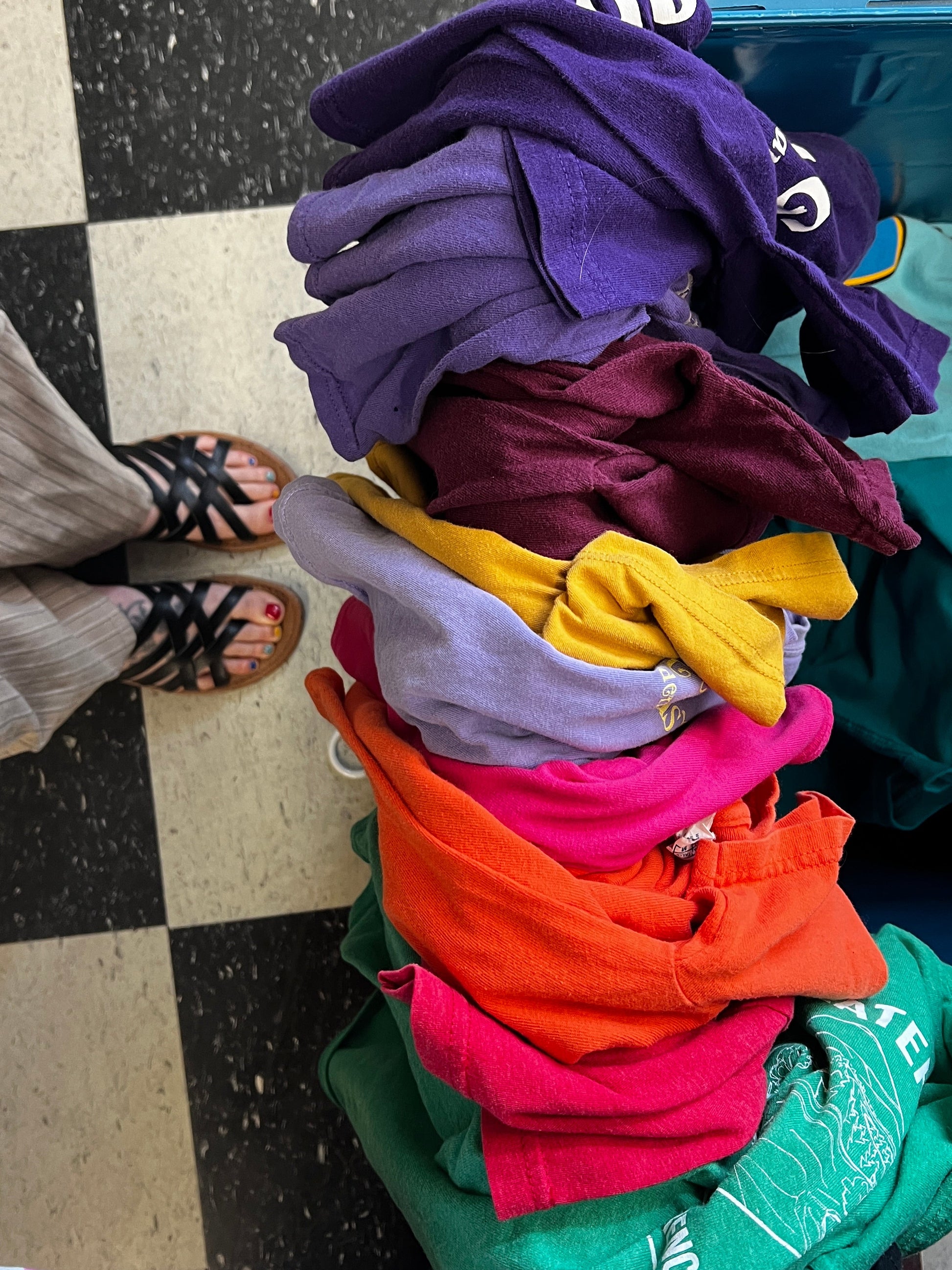 Colorful tshirts stacked on a bin at a thrift store  with a person's feet visible at the bottom.