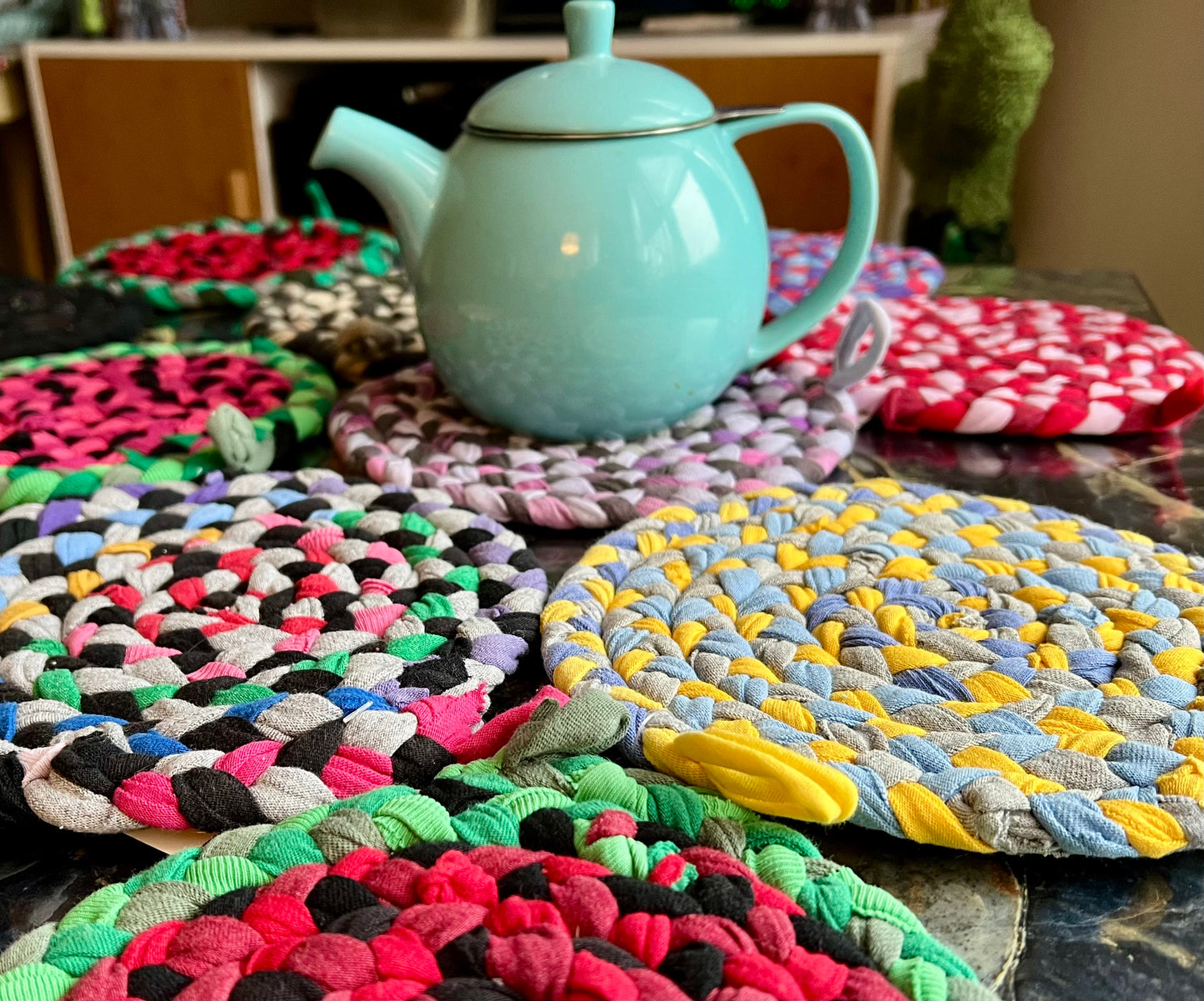 Colorful braided trivets with a teal teapot on a wooden surface.