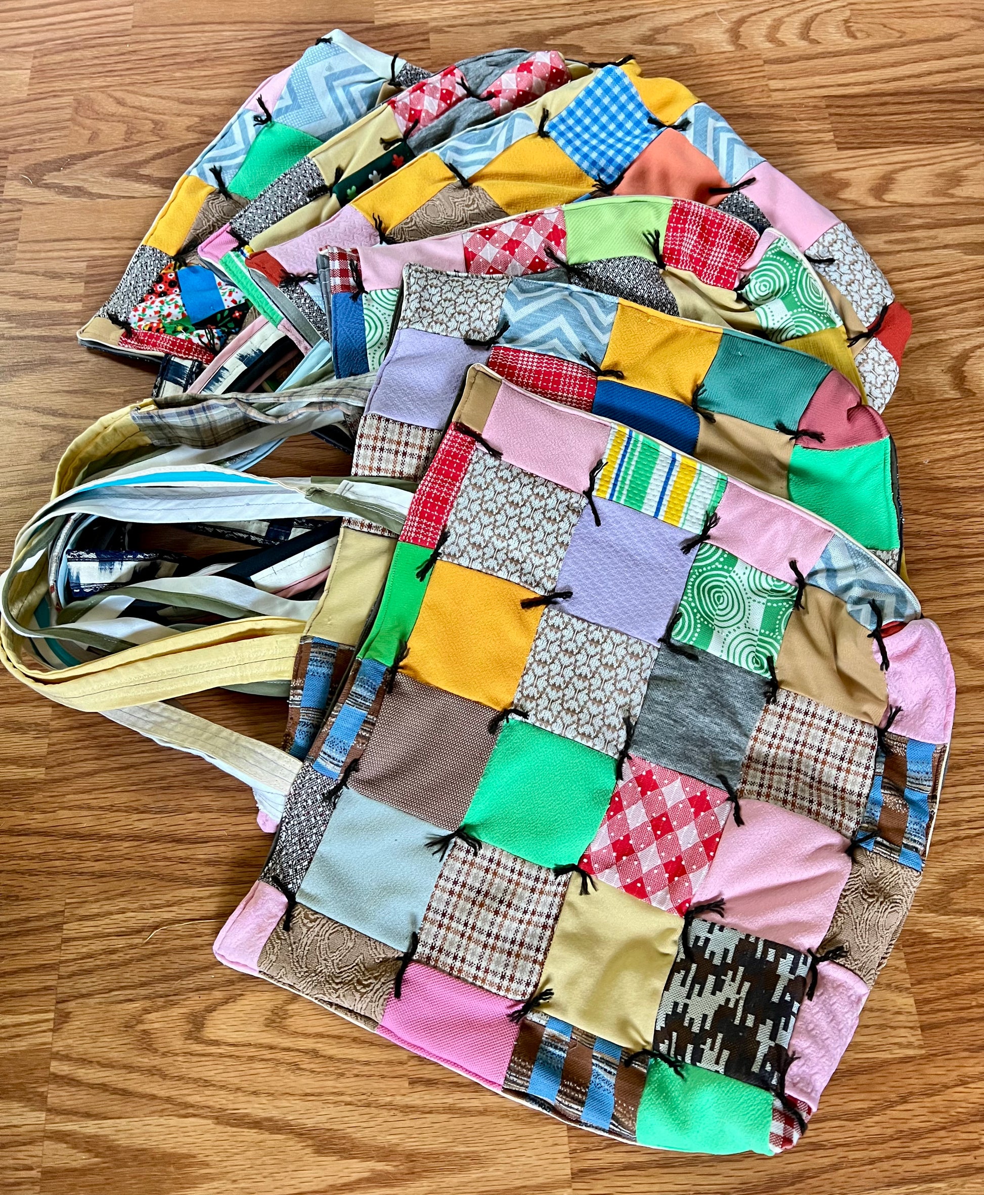A handmade patchwork fabric tote bag with a multicolor quilted pattern, laid flat on a wooden floor. The bag features a variety of patchwork squares in pink, yellow, green, blue, and brown, with a light beige shoulder strap.