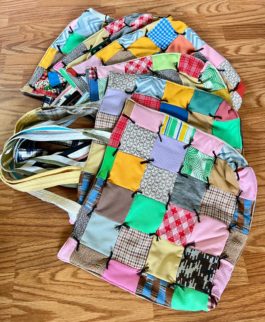 A handmade patchwork fabric tote bag with a multicolor quilted pattern, laid flat on a wooden floor. The bag features a variety of patchwork squares in pink, yellow, green, blue, and brown, with a light beige shoulder strap.