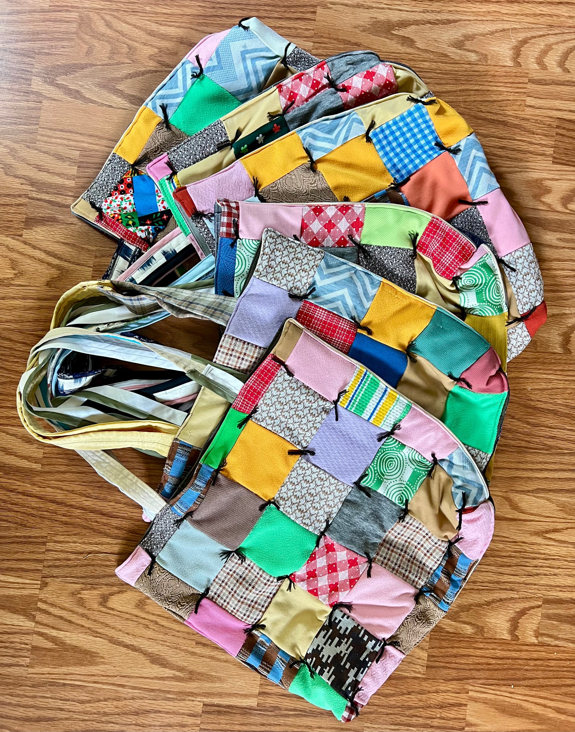 A handmade patchwork fabric tote bag with a multicolor design, laid flat on a wooden floor. The bag features various patchwork squares in pink, yellow, blue, green, and brown, with a striped fabric shoulder strap.