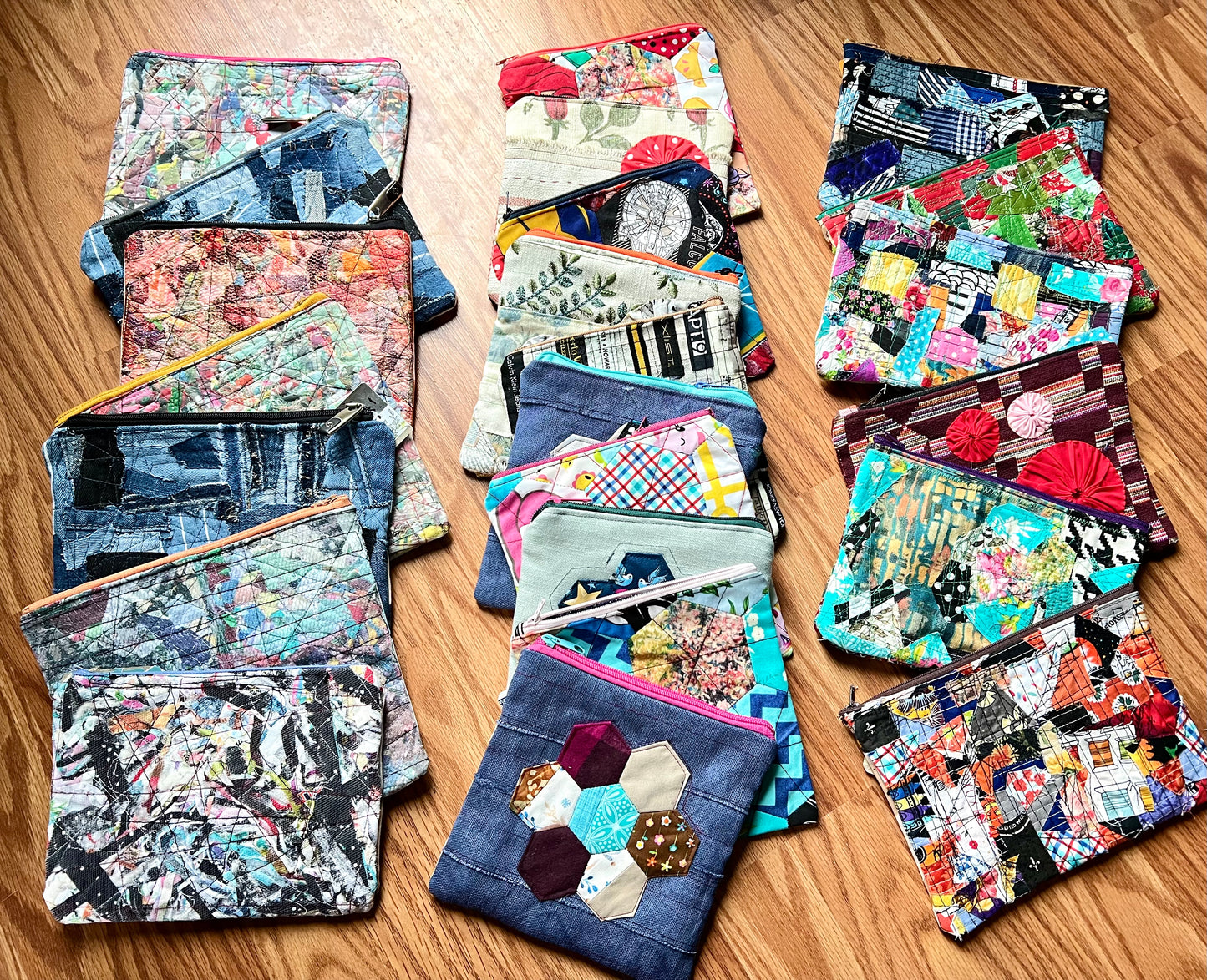 A collection of handmade patchwork zippered pouches made from various colorful fabrics, including denim, floral prints, and abstract patterns, arranged on a wooden surface.