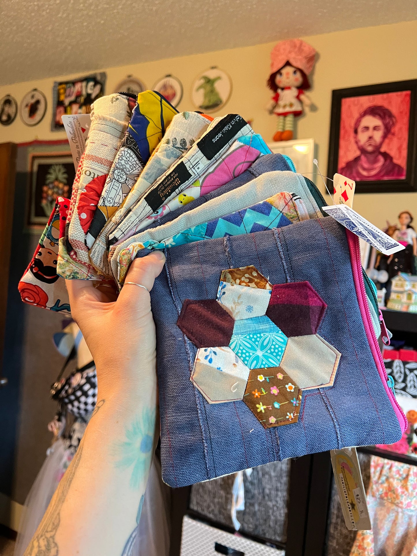 A hand holds a fan of quilted fabric pouches in a studio setting. The front pouch is made of blue denim-like fabric with a hexagonal patchwork design in various colors and patterns.