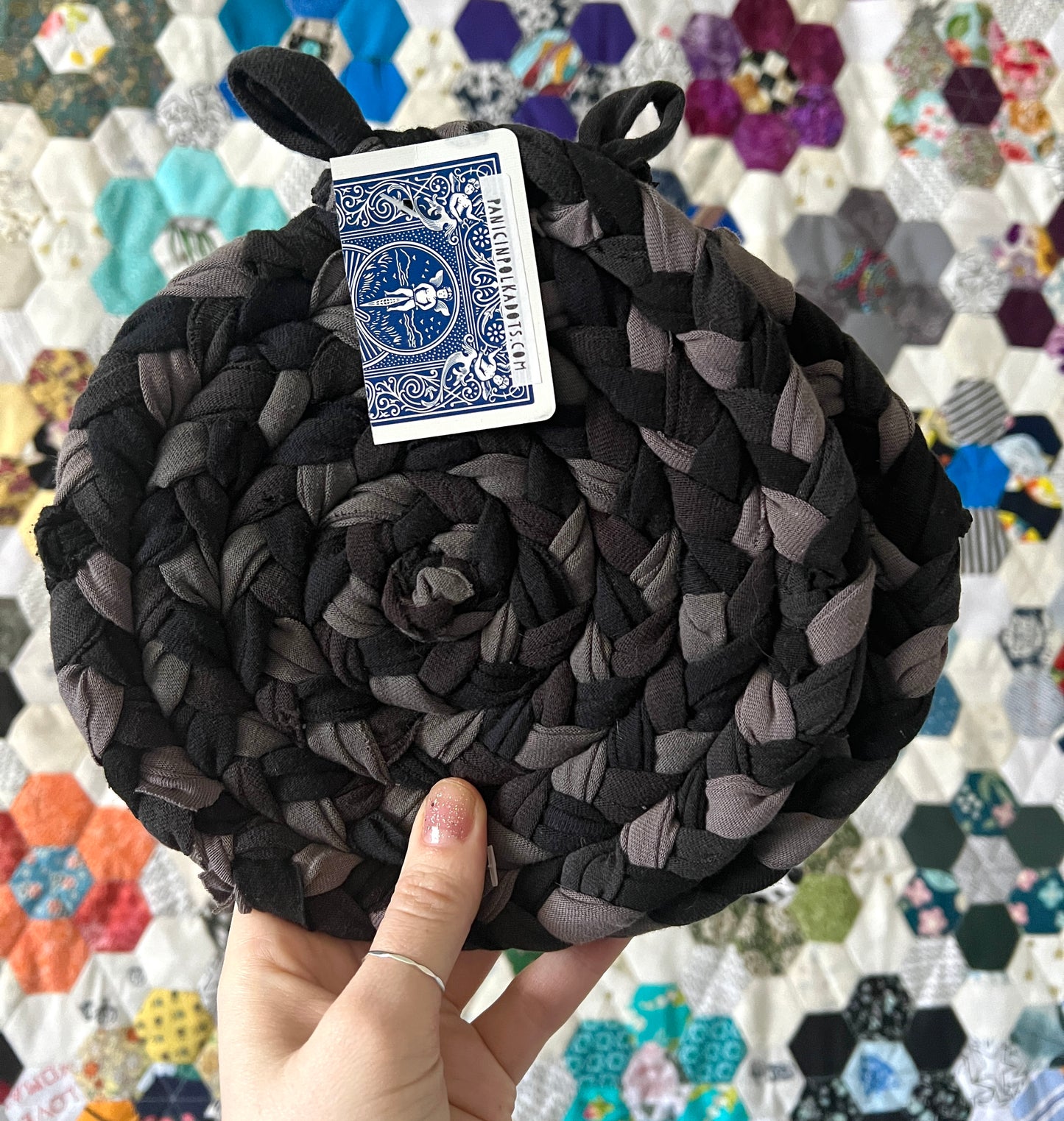 black grey hand braided and hand sewn trivet potholder, held up against a colorful hexie quilt background