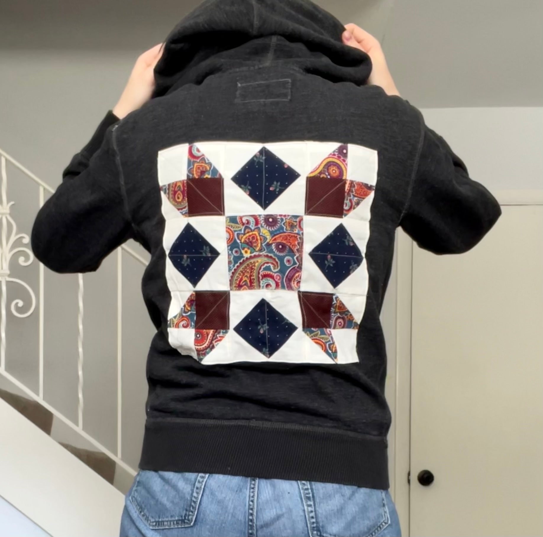 a model wearing blue jeans is showing off the back of this unique hoodie, with arms up holding the hood up