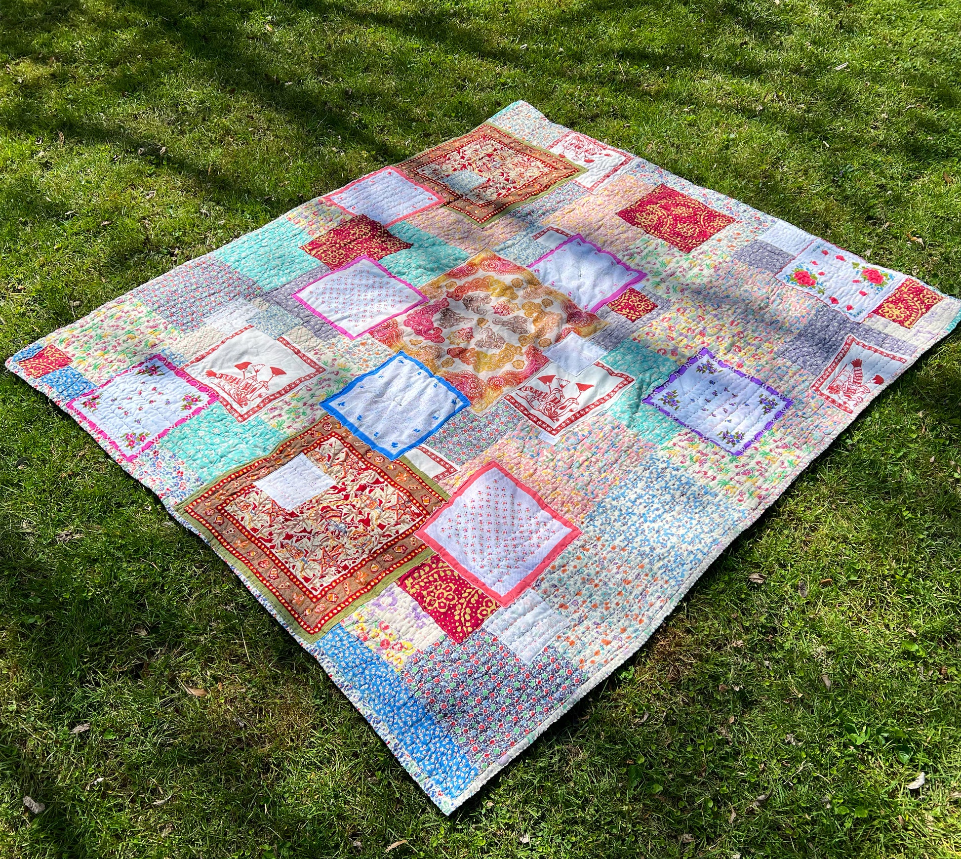a beautiful vintage quilt, which has been mended and completely hand stitched all over, lays on a plush green lawn. Splotches of sunshine dance across the quilt top